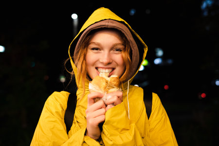 Woman in a yellow hooded raincoat is eating a bun on a city street at nightの写真素材