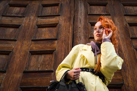 Stylish woman portrait in a yellow oversized raincoat, wearing glasses and carrying a black handbag,...