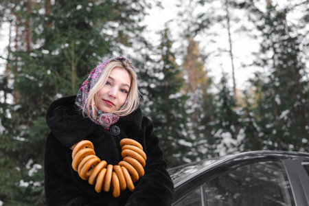 Slavic woman in a black fur coat in the forest poses in a traditional scarf with bagels against the background of a car.の写真素材