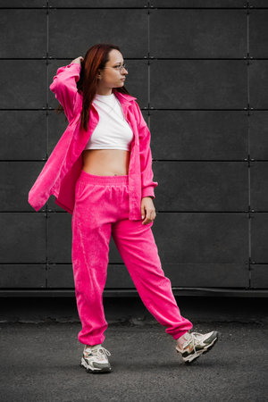 Woman in a pink tracksuit and glasses stands confidently in front of a dark building, showcasing a...