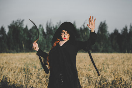 Hooded woman with a scythe stands in a field, exuding a mystical and eerie vibe, suggesting a ritual or witchcraft.の写真素材