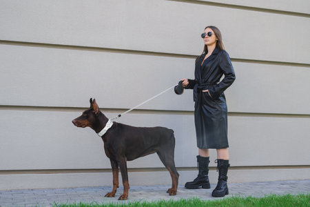 Stylish woman in black leather walks a Doberman on an urban street with modern buildings in the background.の写真素材