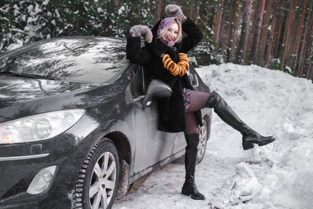 Slavic woman in a black fur coat in the forest poses in a traditional scarf with bagels against the background of a car.の写真素材
