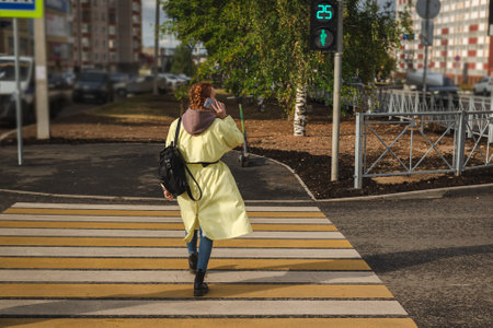 Woman is walking on a crosswalk while talking on her phone, wearing a yellow raincoat, jeans, black boots, and carrying a handbag.の写真素材