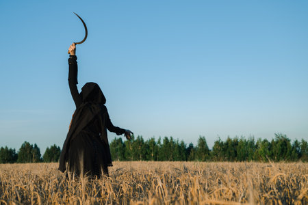 Hooded woman with a scythe stands in a field, exuding a mystical and eerie vibe, suggesting a ritual or witchcraft.の写真素材