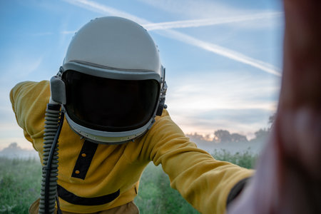 An astronaut in a yellow suit reaches toward the camera in a grassy field, wearing a helmet with a reflective visor, under a sky streaked with contrails.の写真素材