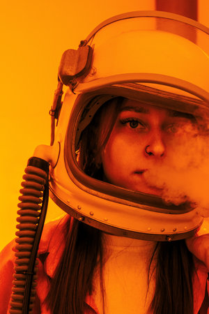 Woman in an astronaut helmet, with smoke emerging, stands against a bright orange background,...