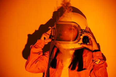 Woman in an astronaut helmet, with smoke emerging, stands against a bright orange background,...