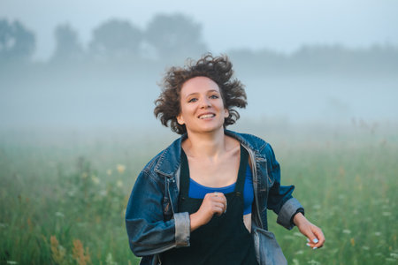 Curly haired 30 year old woman in a sundress walks in the morning misty field.の写真素材