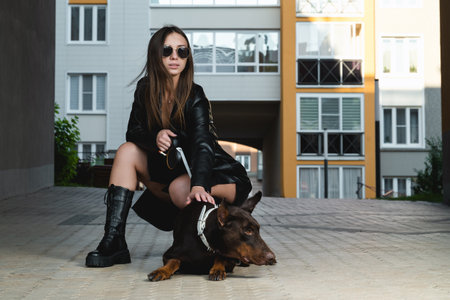 Stylish woman in black leather walks a Doberman on an urban street with modern buildings in the background.の写真素材