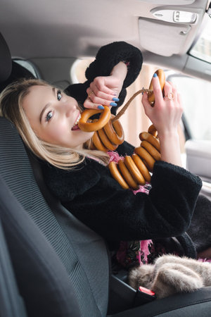 Smiling woman in fur coat in a car enjoys a bagel while wearing a traditional pink scarf and a bagel necklace.の写真素材