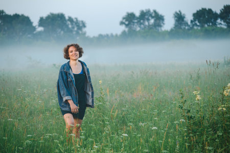 Curly haired woman in a sundress walks in the morning misty field.の写真素材