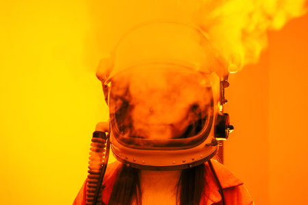 Woman in an astronaut helmet, with smoke emerging, stands against a bright orange background, evoking space exploration and mystery.の写真素材