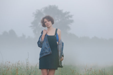 Curly haired woman in a sundress stands in the morning misty field.の写真素材