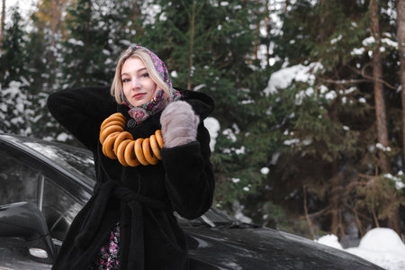 Slavic woman in a black fur coat in the forest poses in a traditional scarf with bagels against the background of a car.の写真素材