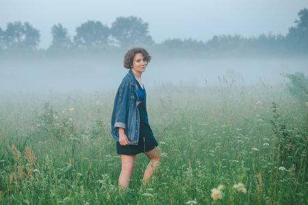 Curly haired 30 year old woman in a sundress walks in the morning misty field.の写真素材