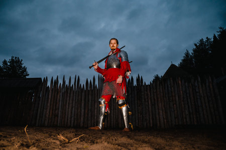 Medieval mustachioed knight in armor with sword, standing near wooden castle at dusk, historical reenactment.の写真素材