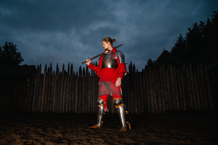 Medieval mustachioed knight in armor with sword, standing near wooden castle at dusk, historical reenactment.の写真素材