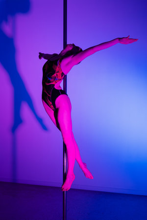 Acrobatic pole dance. Grace and strength, professional dancer. A woman is practicing gymnastics on a pole in the neon light of the studio.の写真素材