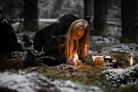 Blonde witch in a black coarse cassock sits on moss in a snowy forest and conjures.の写真素材