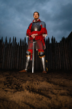 Medieval mustachioed knight in armor with sword, standing near wooden castle at dusk, historical reenactment.の写真素材