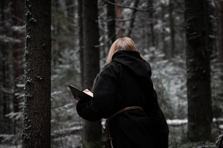 Blonde witch in a black coarse cassock sits on moss in a snowy forest and conjures.の写真素材