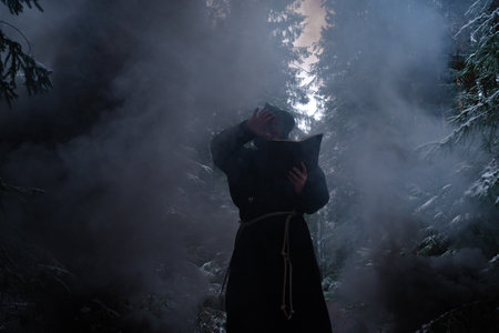 Sorcerer in cloak black is conjuring in a winter forest against a background of thick smoke.の写真素材