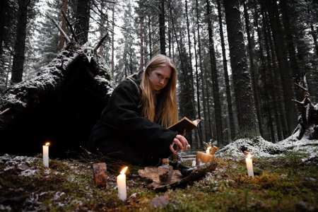 Blonde witch in a black coarse cassock sits on moss in a snowy forest and conjures.の写真素材