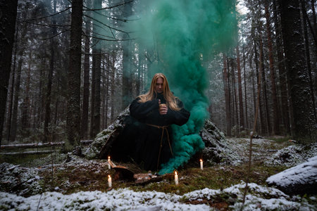 Blonde witch in a black coarse cassock sits on moss in a snowy forest and conjures.