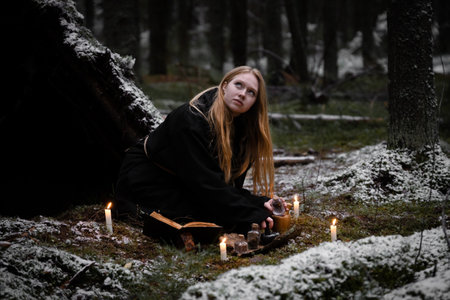 Blonde witch in a black coarse cassock sits on moss in a snowy forest and conjures.の写真素材