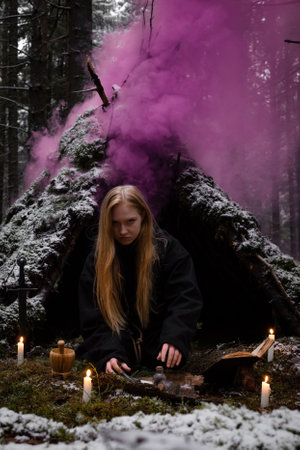 Blonde witch in a black coarse cassock sits on moss in a snowy forest and conjures.の写真素材