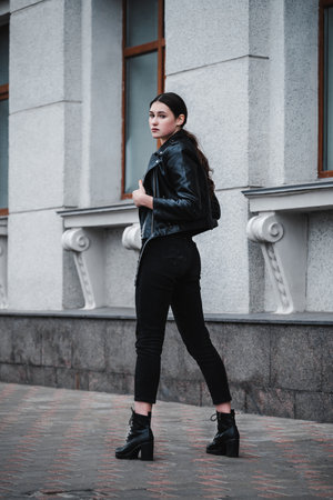 Young woman in a black leather jacket and boots posing confidently in an urban setting.の写真素材