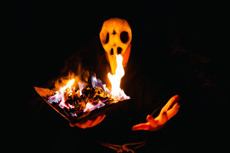 Witch in skull mask a black coarse cassock sits in the night forest and conjures with a burning...