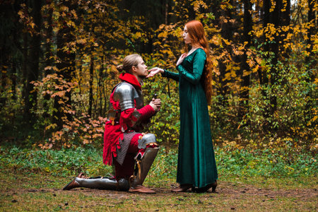 Knight kneeling before a lady in a green dress in medieval autumn forest. Dedication oath in knights.の写真素材