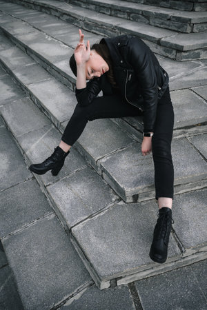 Young woman in a black leather jacket and boots sitting on steps.の写真素材