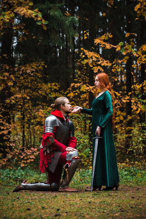 Knight kneeling before a lady in a green dress in medieval autumn forest. Dedication oath in knights.の写真素材