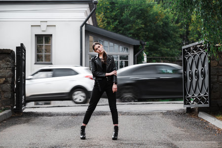 Young woman in a black leather jacket and boots posing confidently in an urban setting.の写真素材