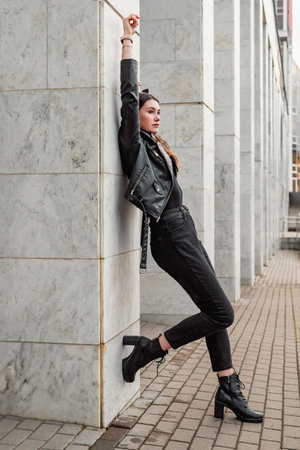 Young woman in a black leather jacket and boots posing confidently in an urban setting.の写真素材