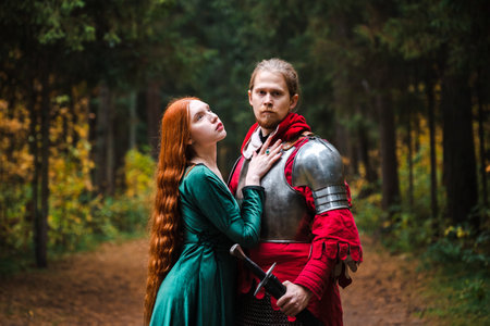 Medieval knight in armor and princess in green dress embrace in a forest setting.の写真素材