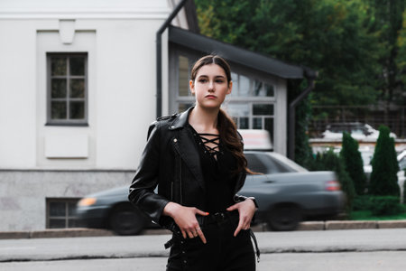 Young woman in a black leather jacket and boots posing confidently in an urban setting.の写真素材
