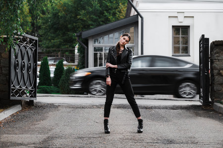 Young woman in a black leather jacket and boots posing confidently in an urban setting.の写真素材