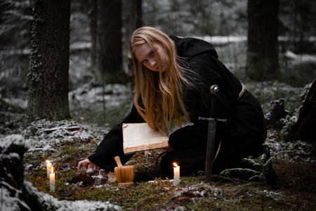 Blonde witch in a black coarse cassock sits on moss in a snowy forest and conjures.の写真素材