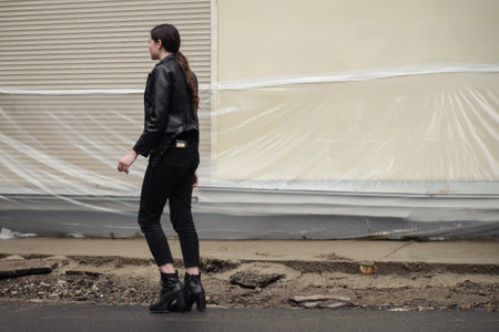 Young woman in a black leather jacket and boots posing confidently in an urban setting.の写真素材