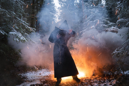 Sorcerer in cloak black is conjuring in a winter forest against a background of thick smoke.の写真素材