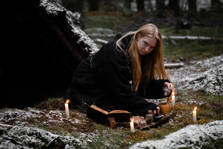 Blonde witch in a black coarse cassock sits on moss in a snowy forest and conjures.の写真素材
