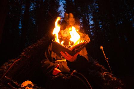 Blonde witch in a black coarse cassock sits in the night forest and conjures with a burning book.の写真素材