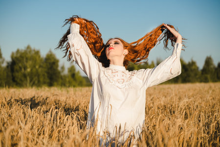 A red-haired Slavic girl in a traditional white outfit rests in a wheat field during harvest.の写真素材