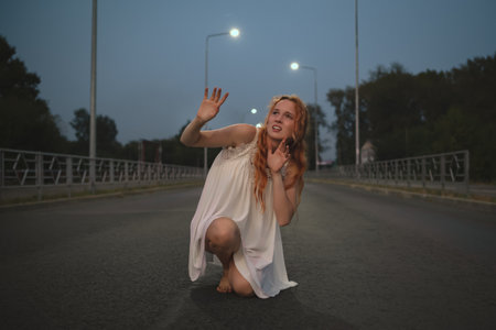 Scared woman in a white dress on a night street, dramatic pose, streetlights.の写真素材