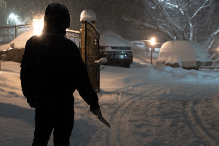 A male stalker or maniac in black clothes with a knife stands on a winter street at the gate of a parking.の写真素材