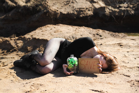 A woman lying on sand holds a âSaveâ sign and a glass jar with greenery, symbolizing environmental awareness.の写真素材